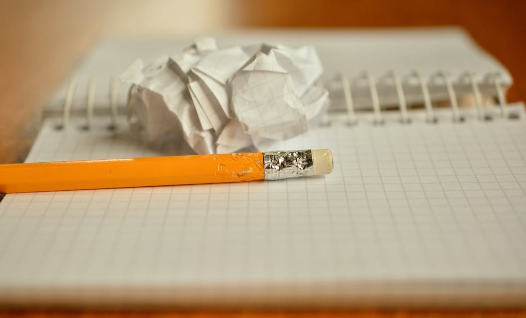 An open, ring-bound notebook with a yellow pencil and a crumpled ball of paper on top of it.