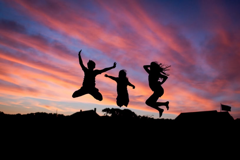 3 people jumping into the air, silhouetted against a pink and purple sunset.